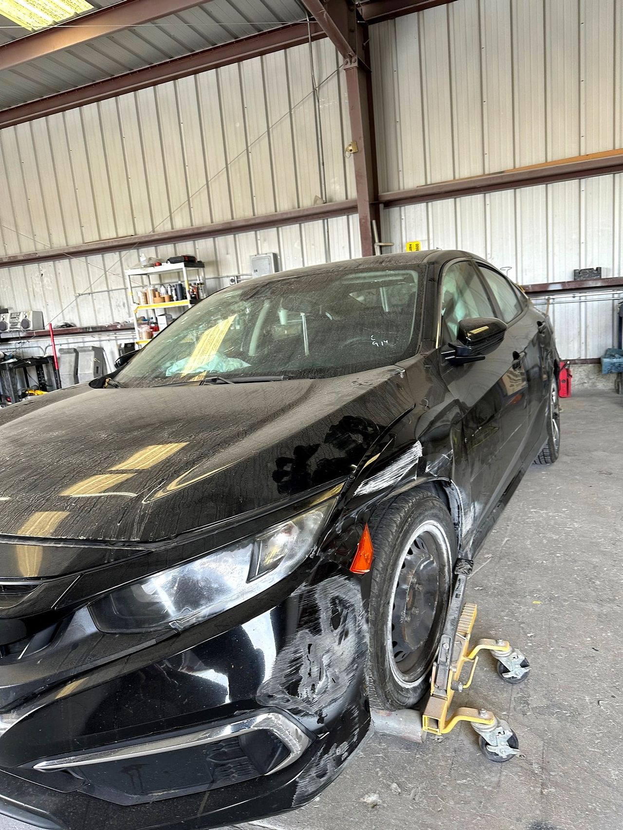 Black Honda Civic with front-end collision damage on wheel dollies inside the shop
