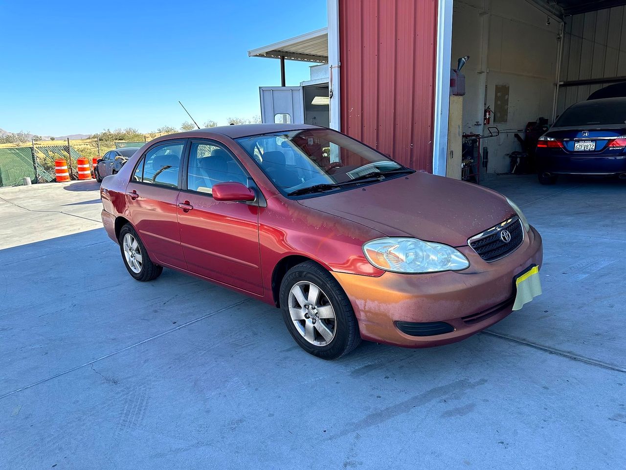 Red Toyota Corolla with sun-peeled clear coat on the hood and bumper outside the open bay