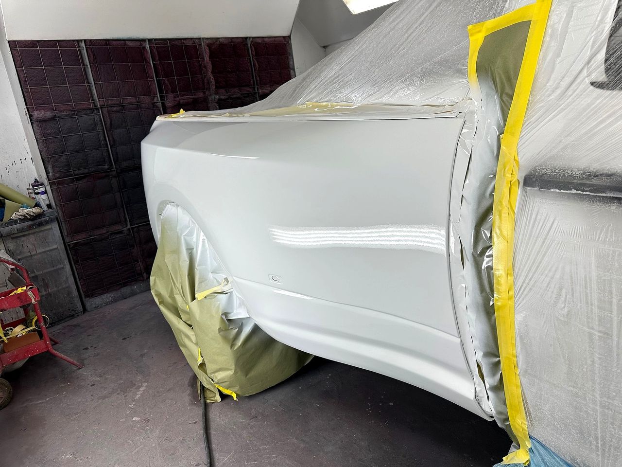 White car's rear quarter panel freshly painted, the rest masked with yellow tape and plastic